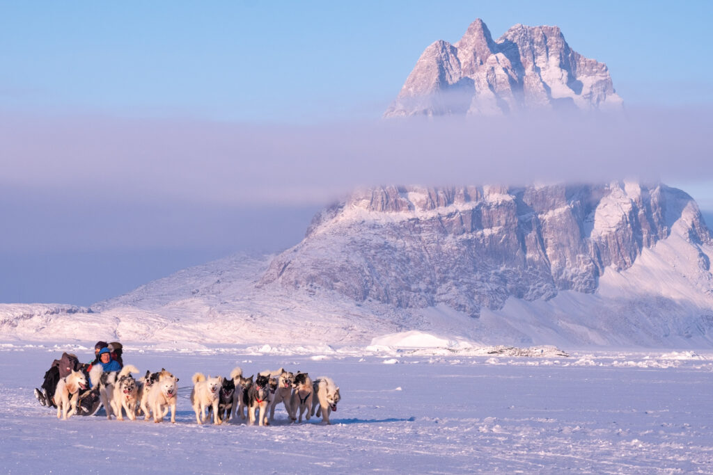 People dogsledding across the sea ice with Uummannaq mountain in the background - Guide to Greenland