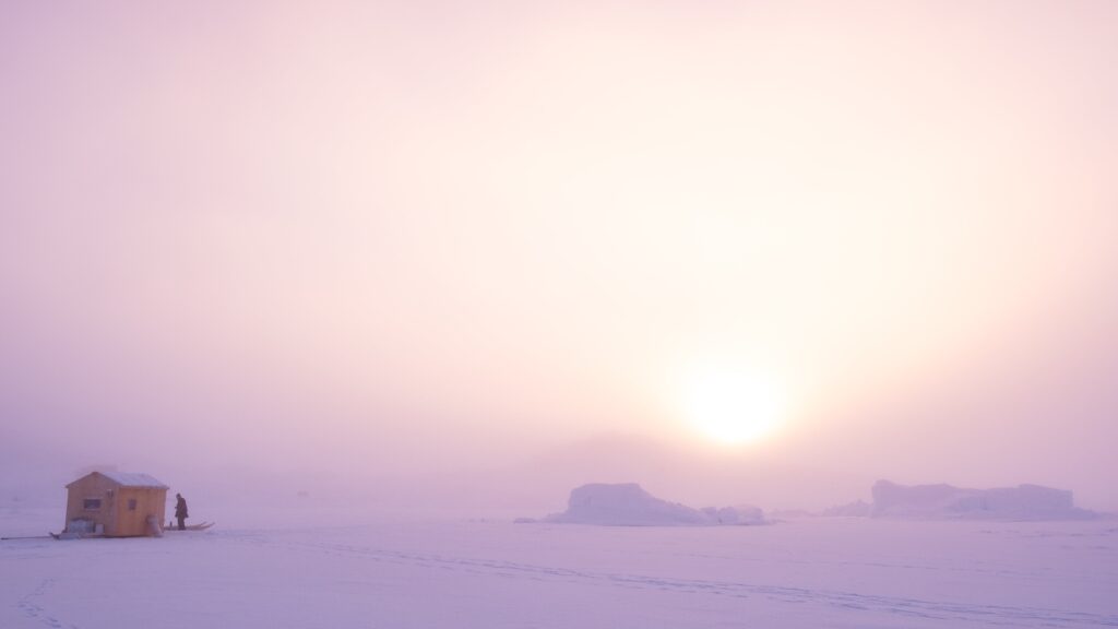 A man in front of a small hut on the sea ice, pink sun in the background